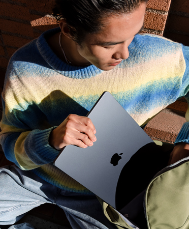 A student sitting outside. He pulls a closed MacBook from his backpack.