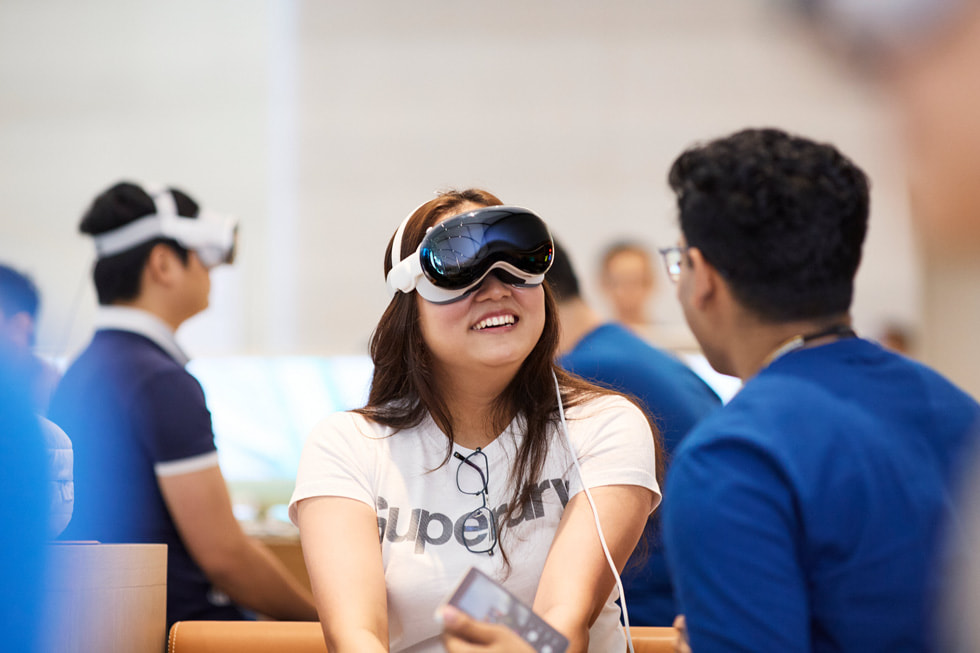 عميلة ترتدي Apple Vision Pro وتبتسم أثناء حديثها مع أحد أعضاء الفريق الجالس بجوارها.