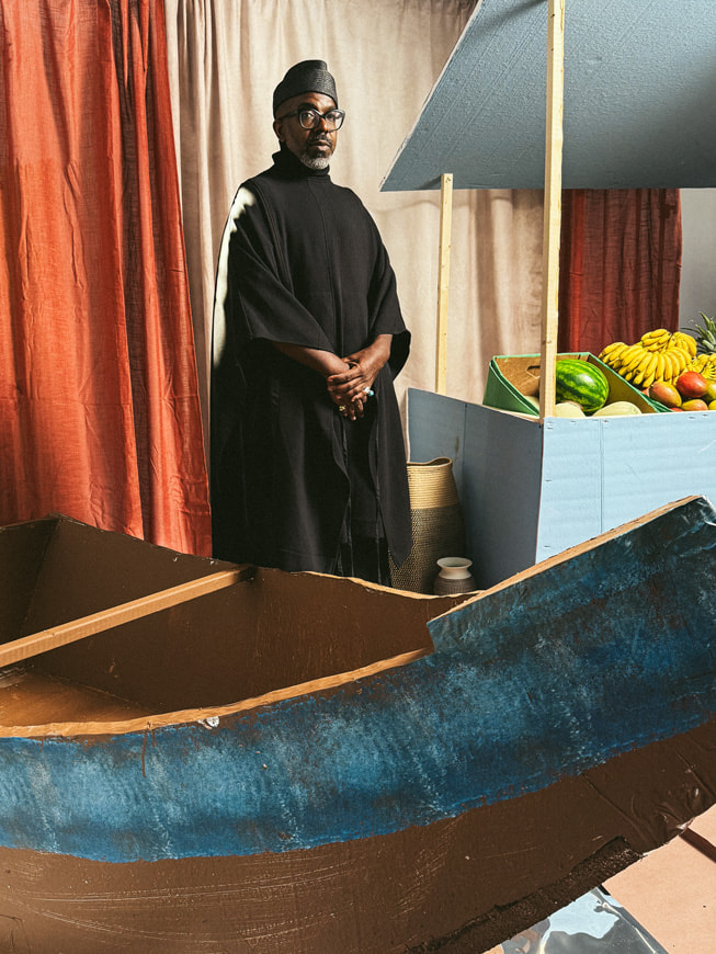 Retrato de Aaron Potts, diseñador de A. Potts, con una túnica negra larga y un sombrero negro, junto a un fondo creado para parecer un puesto de frutas.
