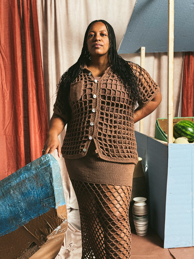 Retrato de la diseñadora Nia Thomas con una falda marrón de crochet y un top haciendo juego, sobre un fondo creado para parecer un puesto de frutas. Foto tomada con un iPhone 16 Pro Max por Karston Tannis.